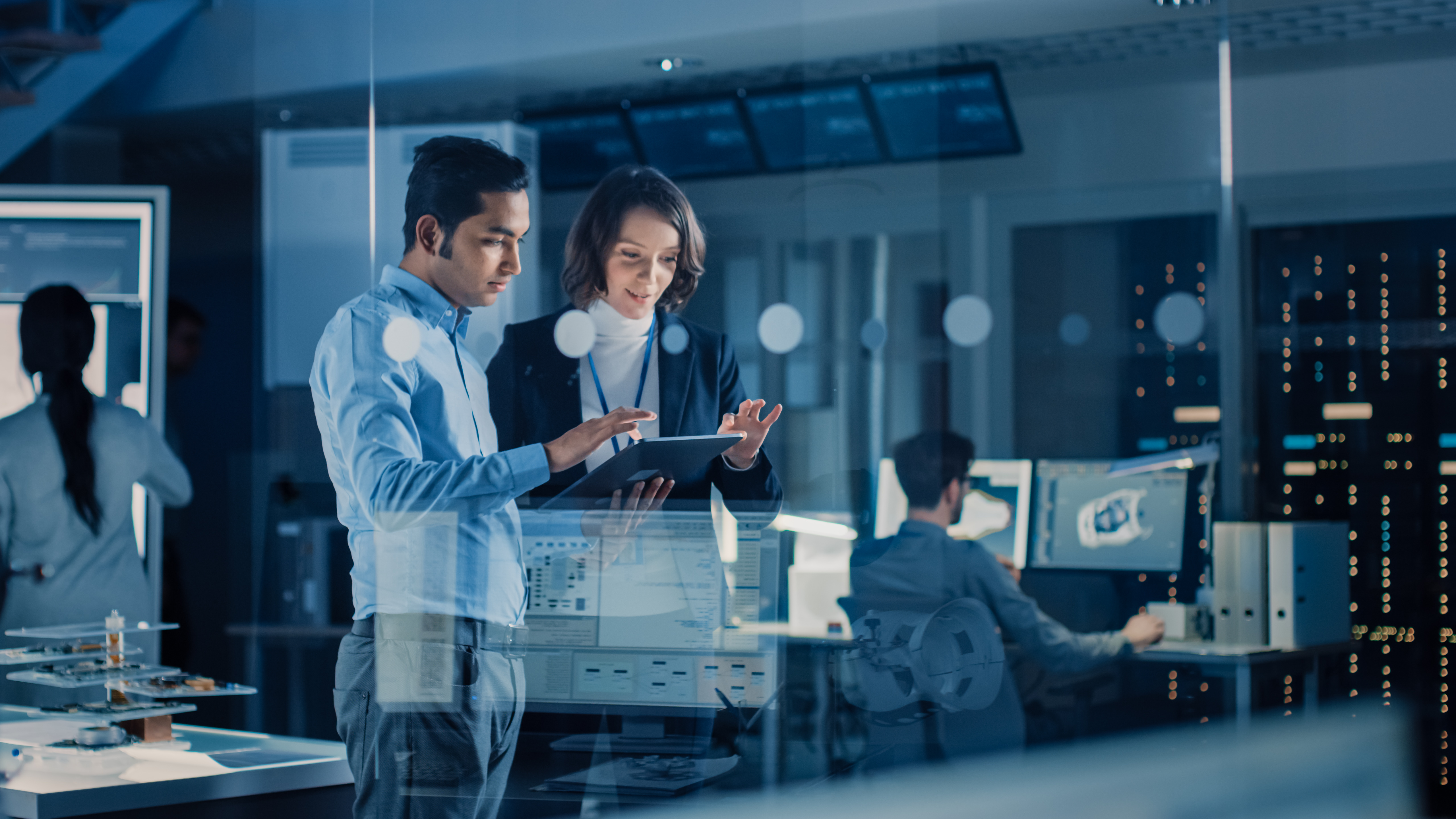 Two technology professionals reviewing data on a tablet in a research facility, representing market analysis amid semiconductor and memory supply constraints.